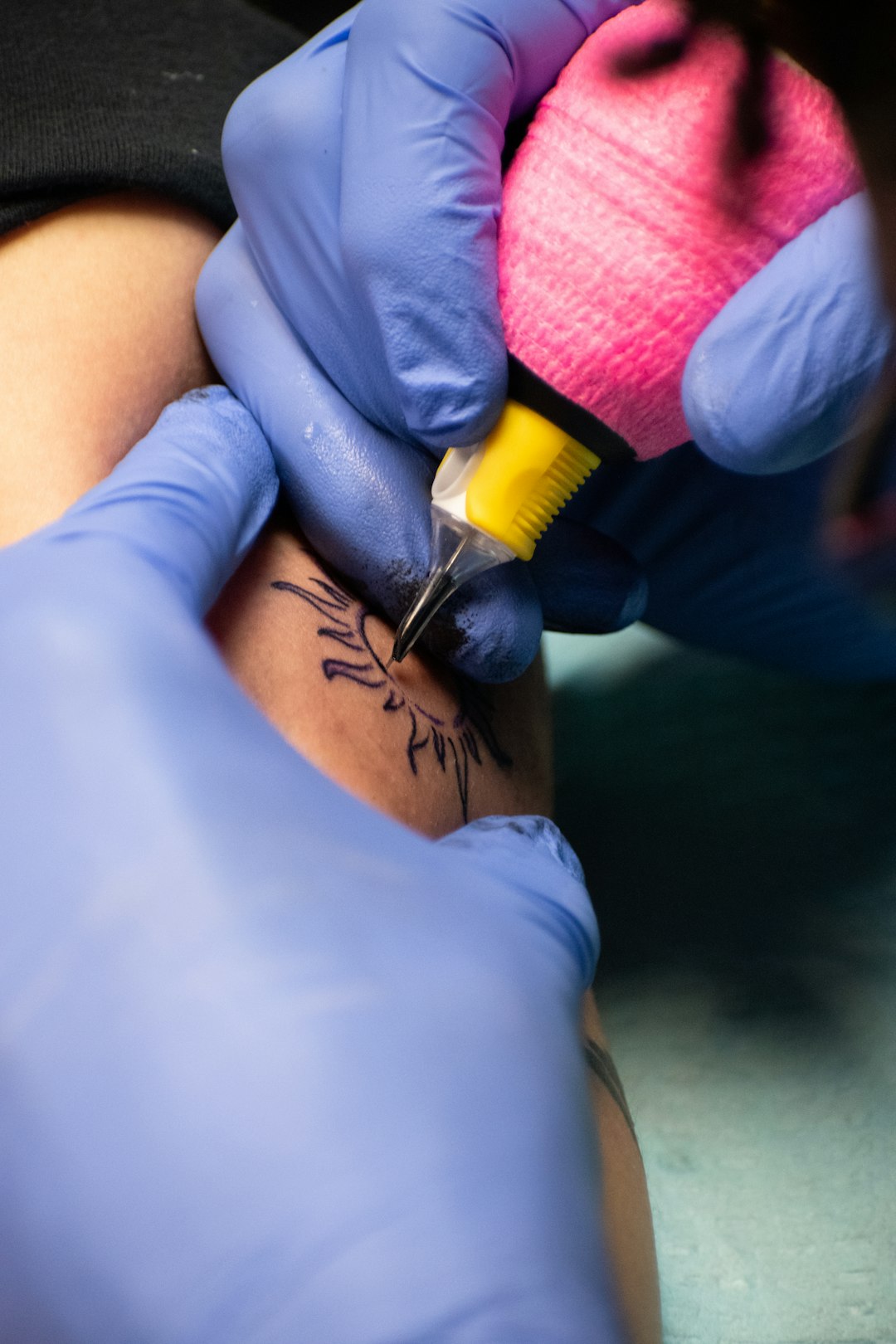 A close up shot shows a tattoo artists gloved hands meticulously working on a detailed line-art tattoo on a persons skin using a pink wrapped tattoo machine.